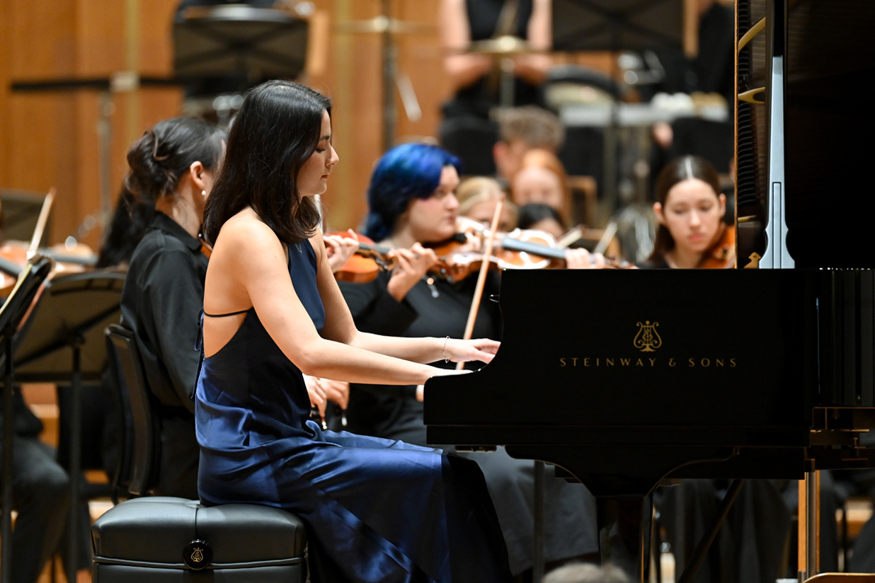 A female student wearing a blue dress, playing the piano, with an orchestra behind her, on a stage